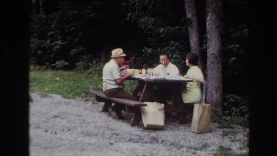 Ottawa 1967 frame with adults seated around a picnic table, food on the tabletop, paper bags on the ground, and dense trees behind them.