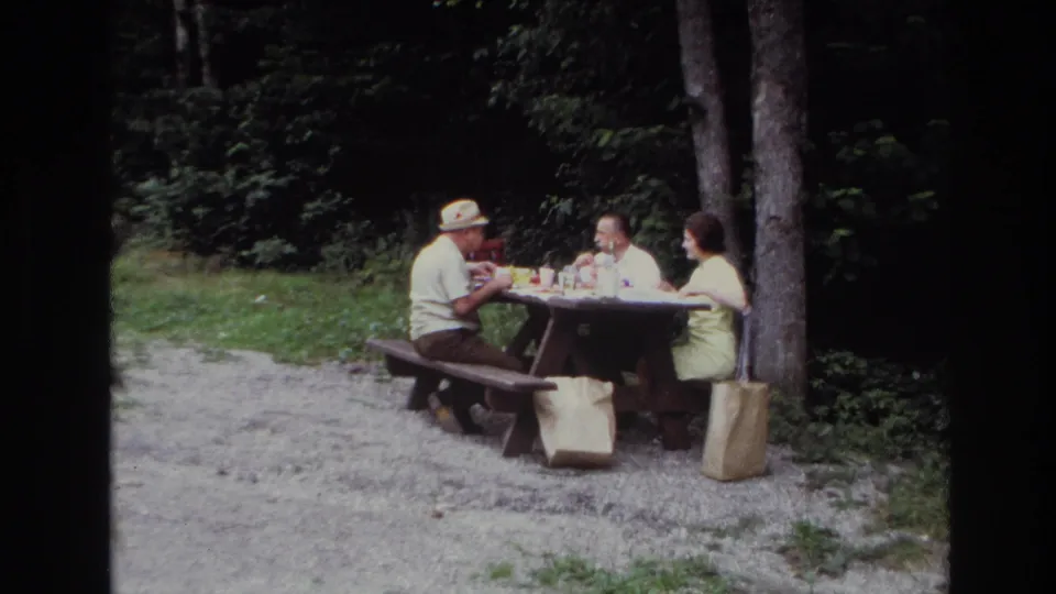 Ottawa 1967 home-movie view of three adults at a wooden picnic table, surrounded by trees, grass, gravel, and picnic bags.