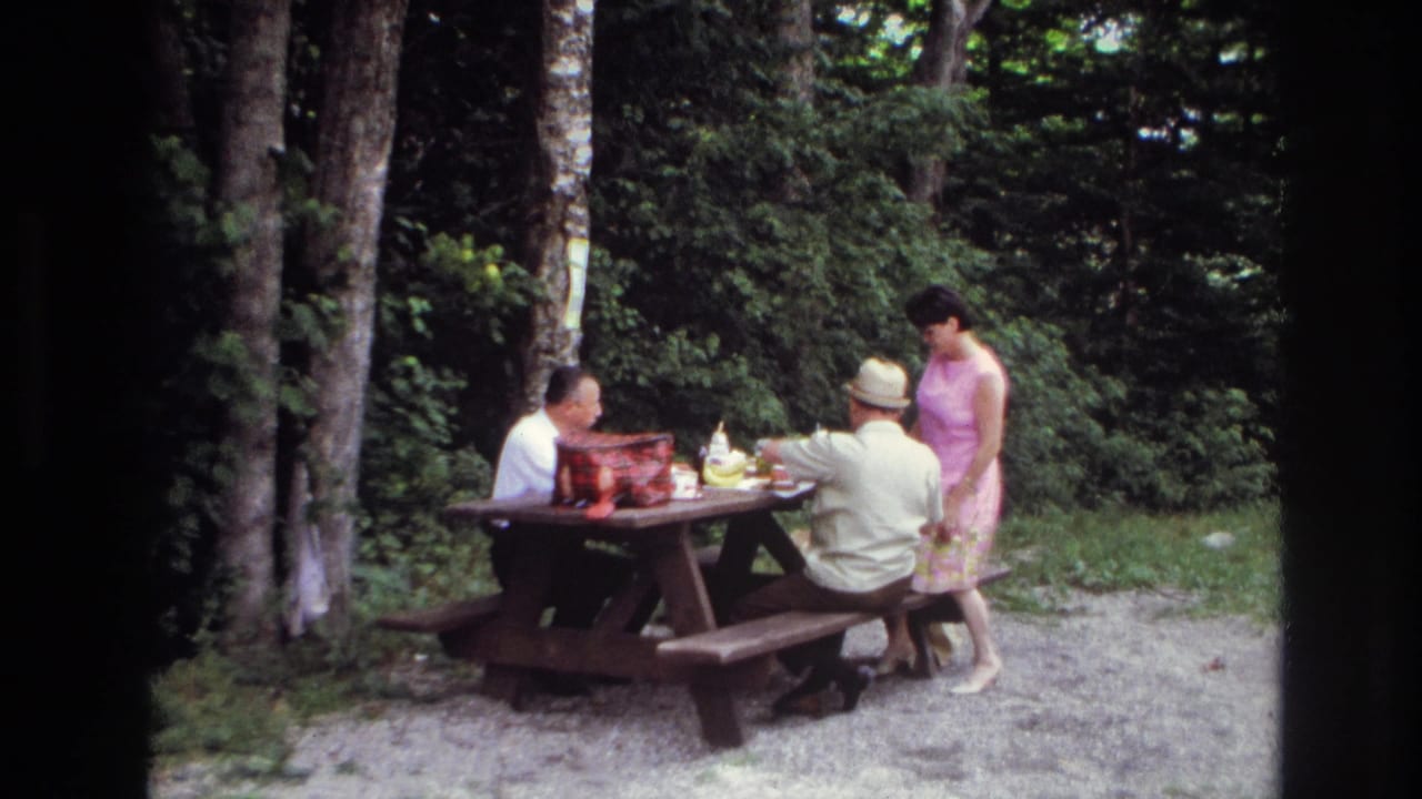 1967 Ottawa home-movie frame showing two men seated at a picnic table while a woman in a pink dress stands beside them.