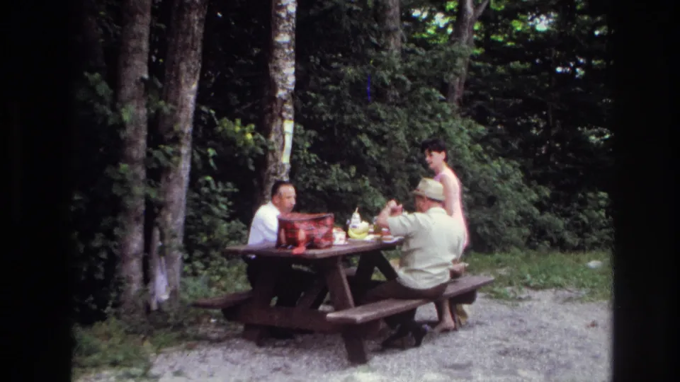 Home-movie frame from Ottawa in 1967 showing adults gathered around a picnic table in a wooded outdoor area.
