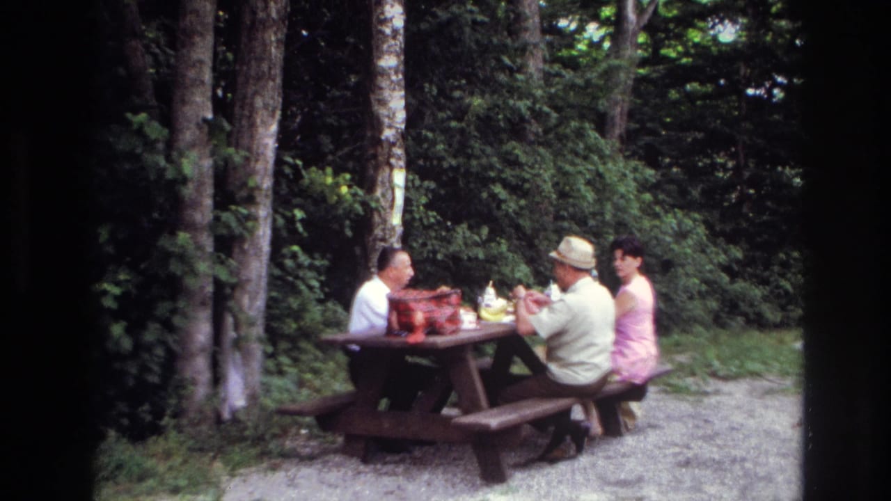 1967 Ottawa home-movie frame of two men and one woman seated at a wooden picnic table in a forest clearing.