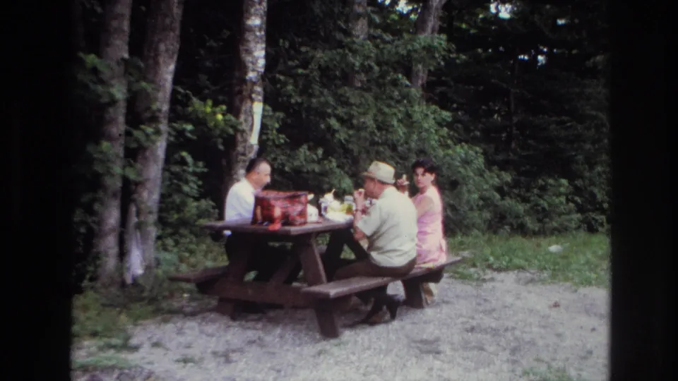 The 1967 Ottawa picnic scene holds on three adults seated together at a table in a wooded clearing.