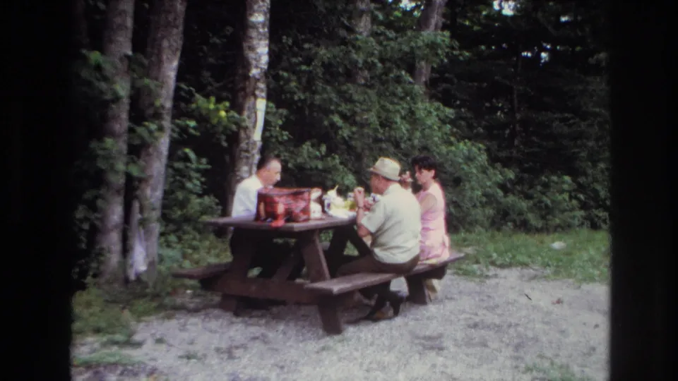 In 1967 Ottawa home-movie footage, three adults appear to eat at a wooden picnic table beside trees and dense green foliage.