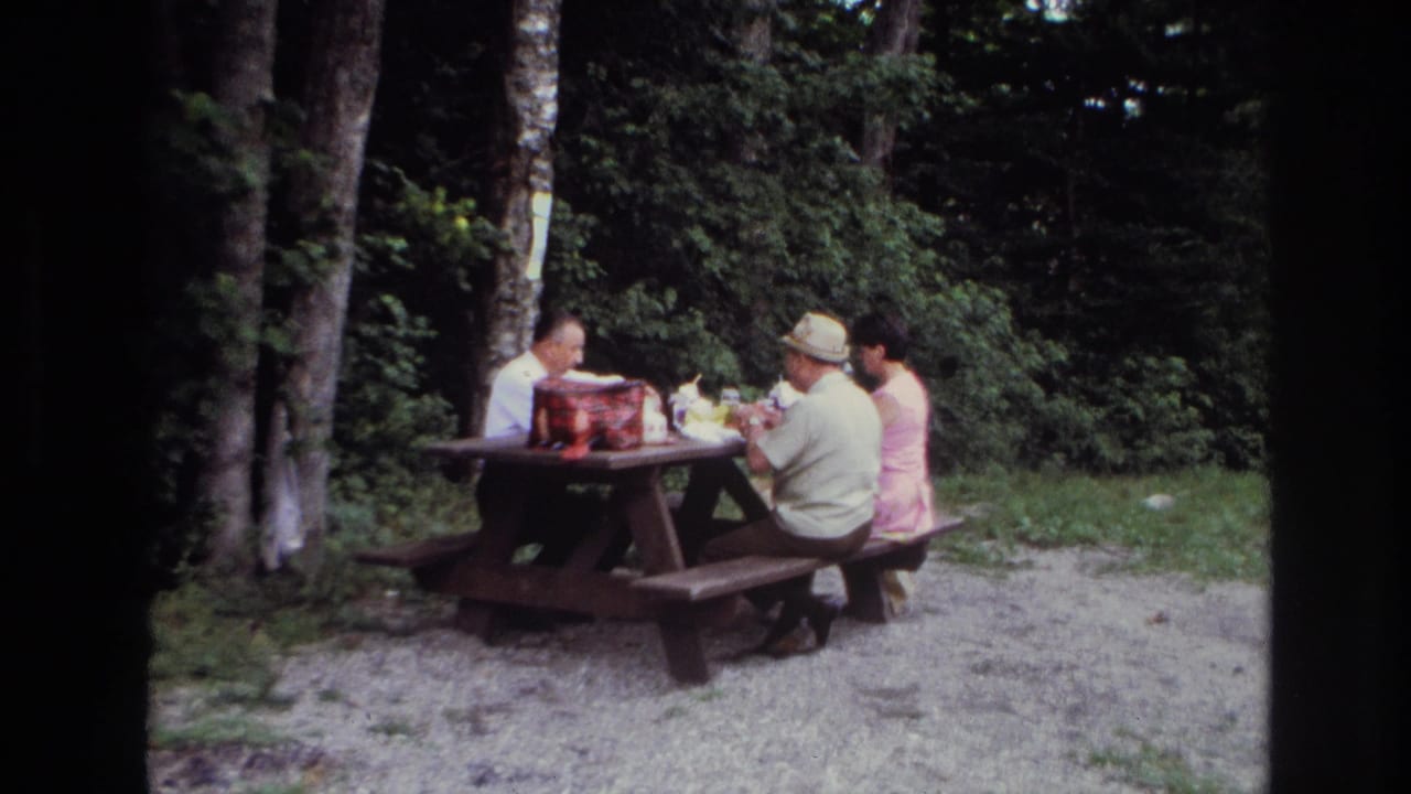 In Ottawa 1967 home-movie footage, three adults sit around a wooden picnic table with food and a red picnic bag nearby.
