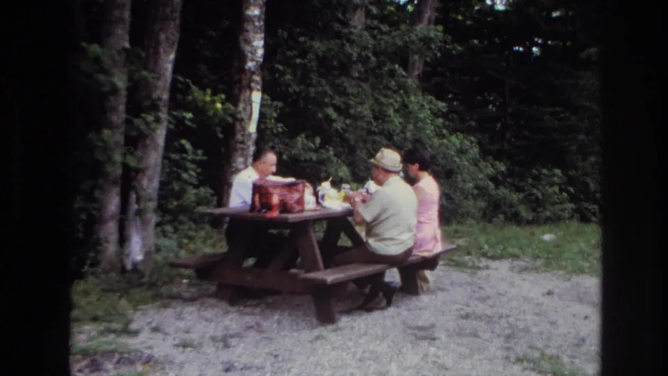 In Ottawa 1967 home-movie footage, three adults sit around a wooden picnic table with food and a red picnic bag nearby.