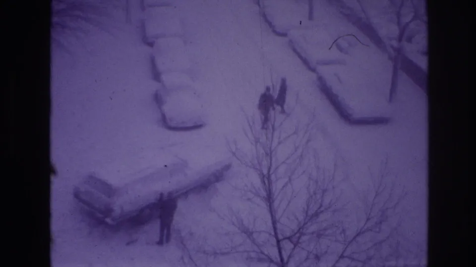 High-angle 1967 Ottawa home-movie frame showing people walking along a snowy street with cars buried in snow and bare tree branches in front.
