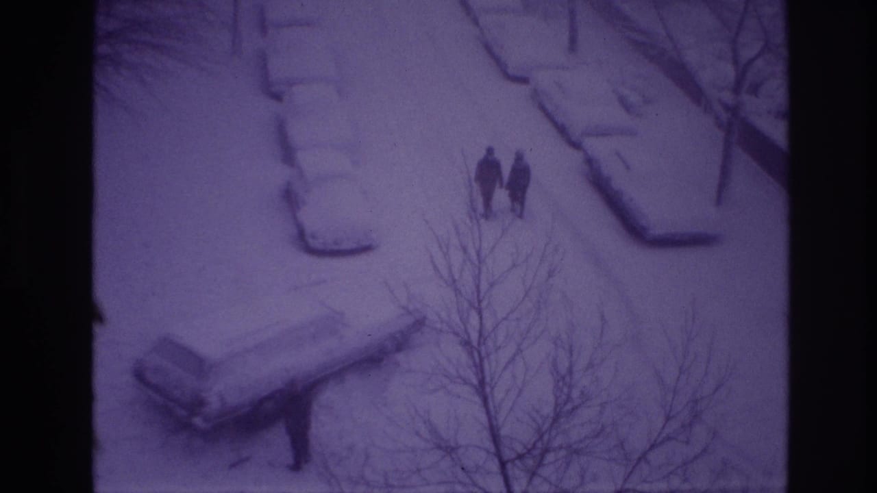 High-angle 1967 Ottawa frame with two bundled pedestrians on a snow-covered road or sidewalk, parked cars under snow, and bare branches nearby.
