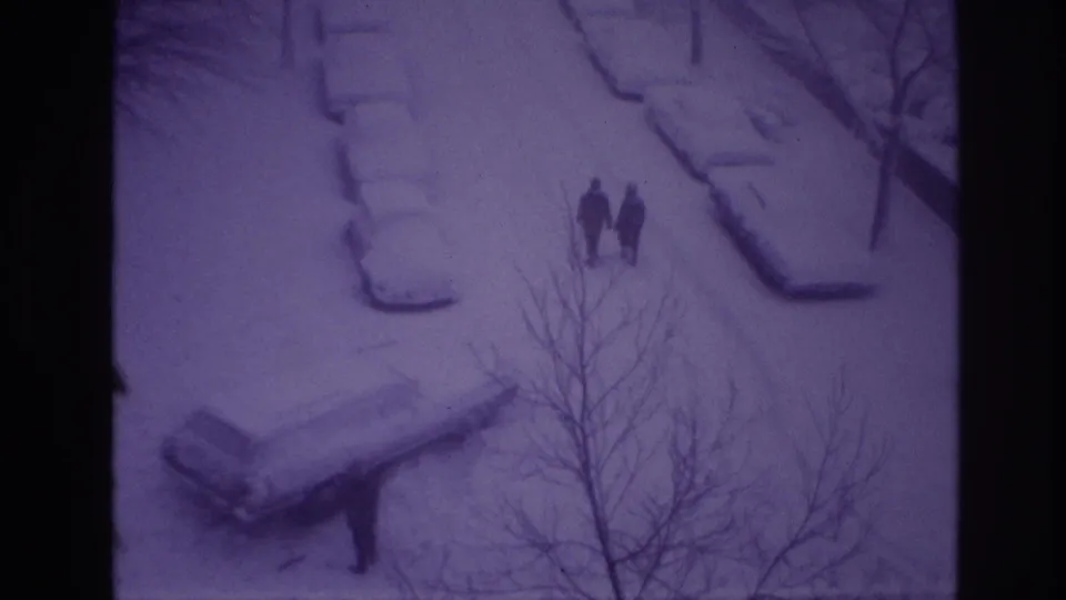 High-angle 1967 Ottawa frame with two bundled pedestrians on a snow-covered road or sidewalk, parked cars under snow, and bare branches nearby.