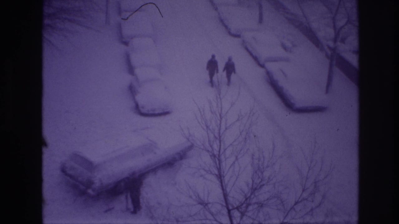 1967 Ottawa home-movie still showing two distant walkers on a snow-covered residential street, with snow-laden parked cars and foreground branches.
