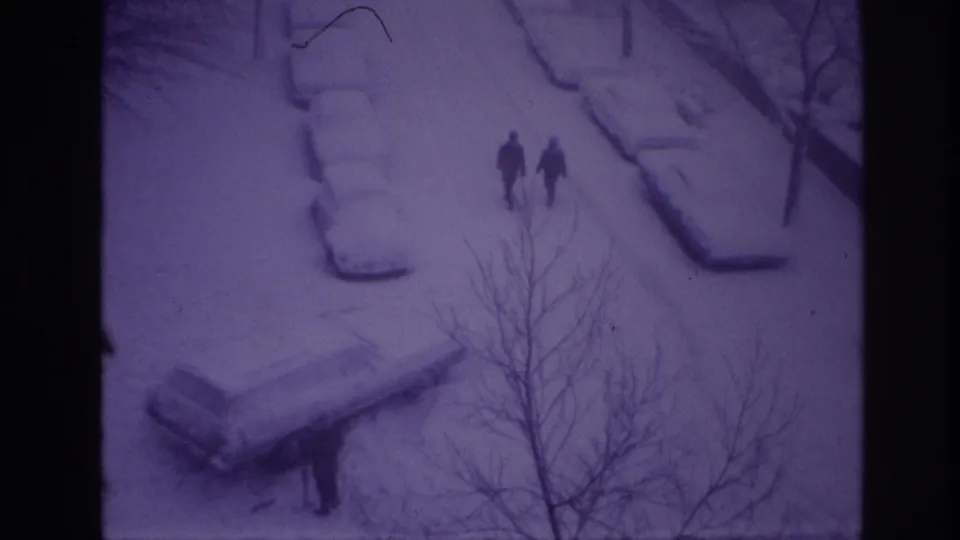 1967 Ottawa home-movie still showing two distant walkers on a snow-covered residential street, with snow-laden parked cars and foreground branches.