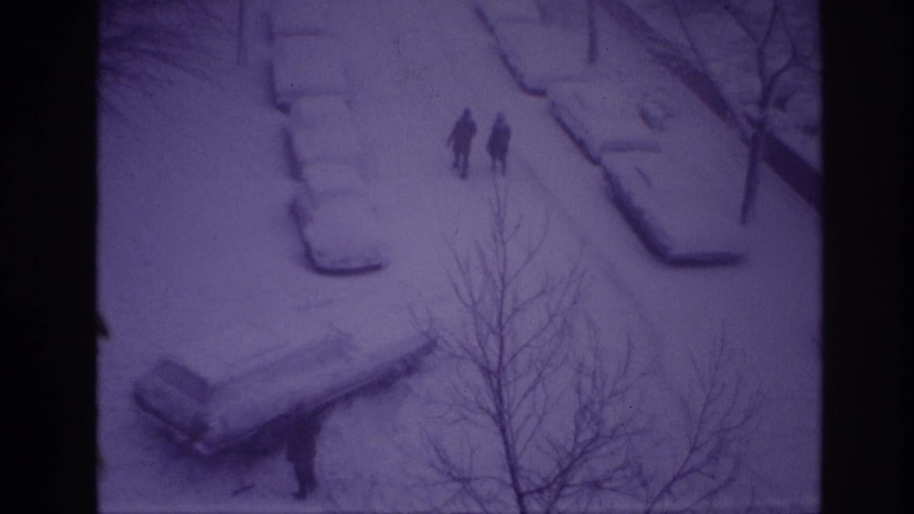 High-angle 1967 Ottawa winter frame with two people walking through a snow-covered street scene, parked cars under snow, and bare branches crossing the view.
