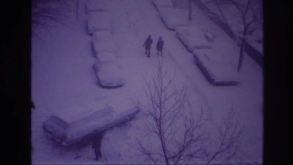 High-angle 1967 Ottawa winter frame with two people walking through a snow-covered street scene, parked cars under snow, and bare branches crossing the view.