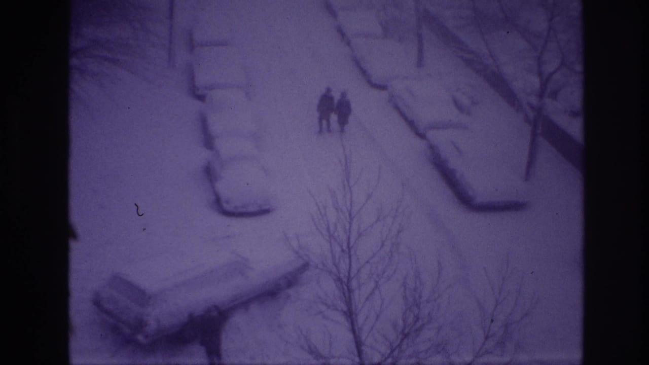 1967 Ottawa home-movie frame showing two distant pedestrians on a snowy street lined with cars partly buried under snow.
