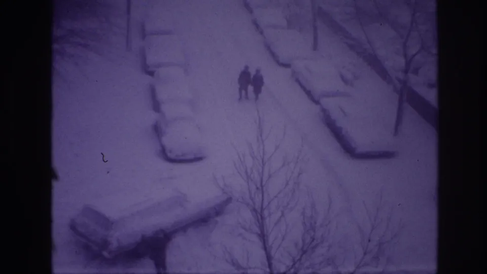 1967 Ottawa home-movie frame showing two distant pedestrians on a snowy street lined with cars partly buried under snow.