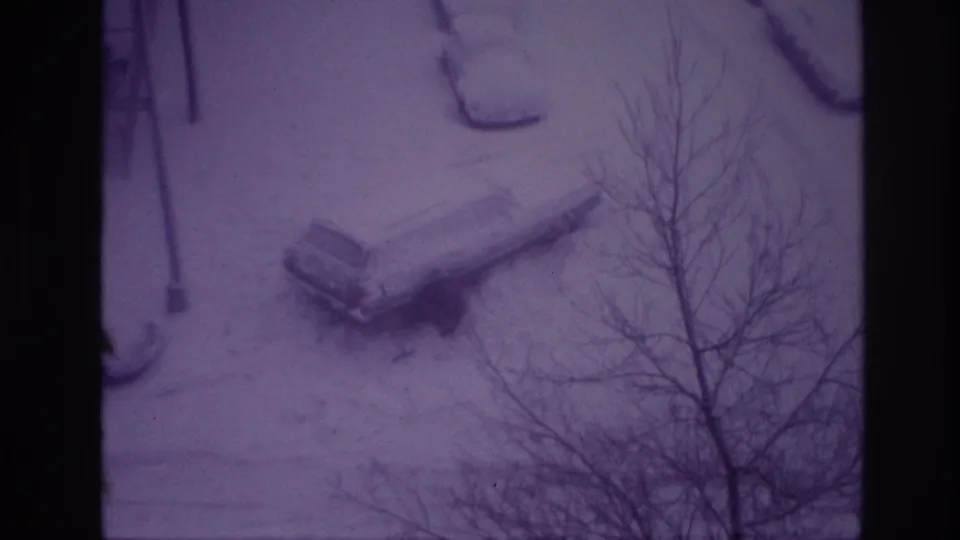 High-angle 1967 Ottawa home-movie frame of a snow-covered parked car and winter-bare trees along a residential street.
