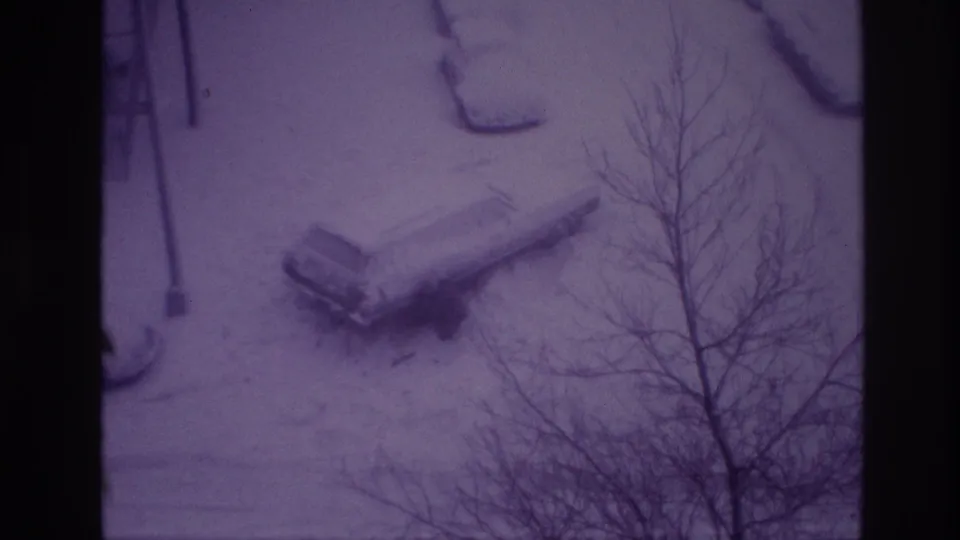 Elevated 1967 Ottawa home-movie view of a parked car buried in snow, with bare branches crossing the frame.