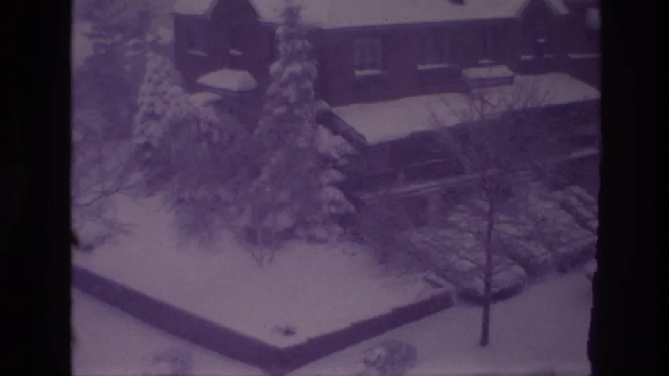 1967 Ottawa home-movie frame of a snowy residential yard, brick house, evergreen trees, parked cars, and walkways.