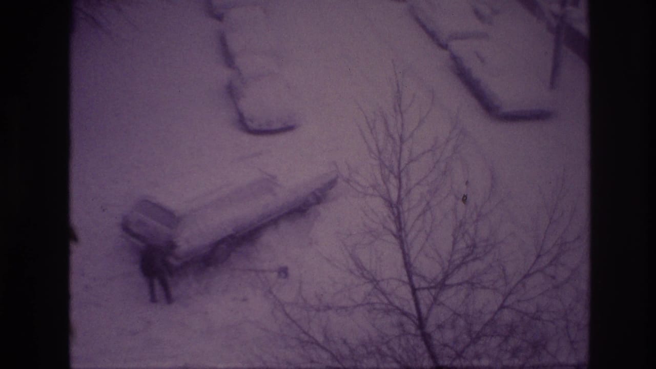 1967 Ottawa home-movie frame showing a car partly buried in snow while a bundled person works near it, viewed through bare branches.