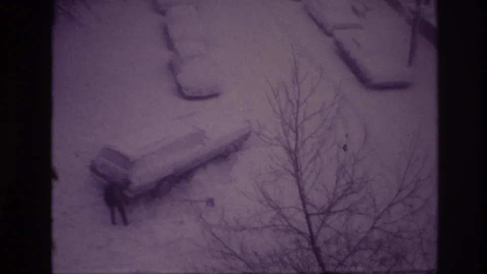 1967 Ottawa home-movie frame showing a car partly buried in snow while a bundled person works near it, viewed through bare branches.