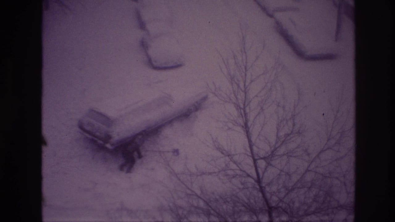 1967 Ottawa 8mm frame showing a person clearing snow around the rear area of a car in a residential driveway.