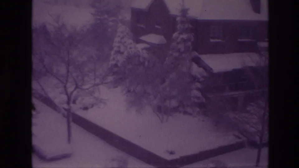 1967 Ottawa home-movie frame widens to a snowy residential area with houses, parked vehicles, leafless trees, and snow-laden evergreens.