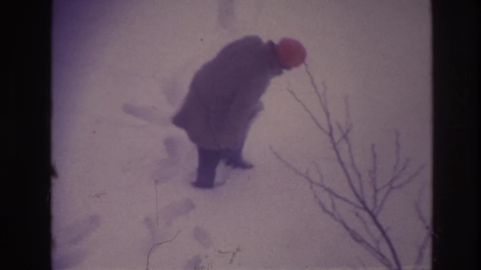 In 1967 Ottawa footage, a bundled person wearing a red hat steps through deep snow near bare foreground branches.