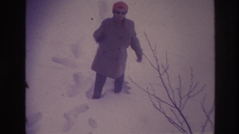 The older woman stands in the snowy Ottawa setting, wearing a long coat, dark gloves, and bright hat as she faces the camera.