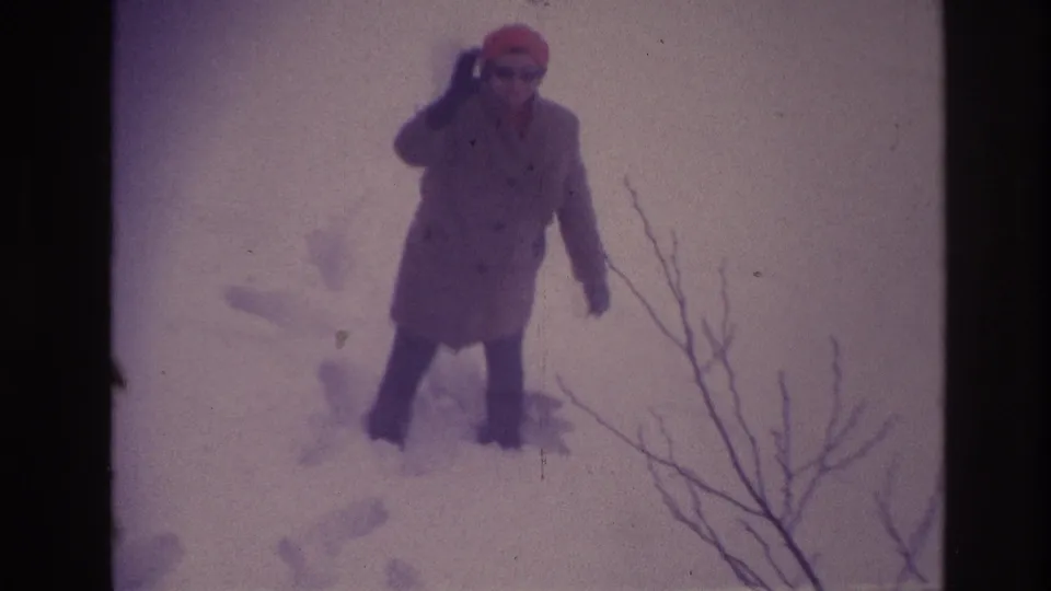 In the 1967 Ottawa home-movie frame, the older woman raises one hand in greeting while standing in deep snow.