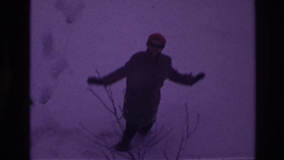The 1967 Ottawa home-movie frame shows the woman moving through deep snow near bare branches, with a dark purple film cast.