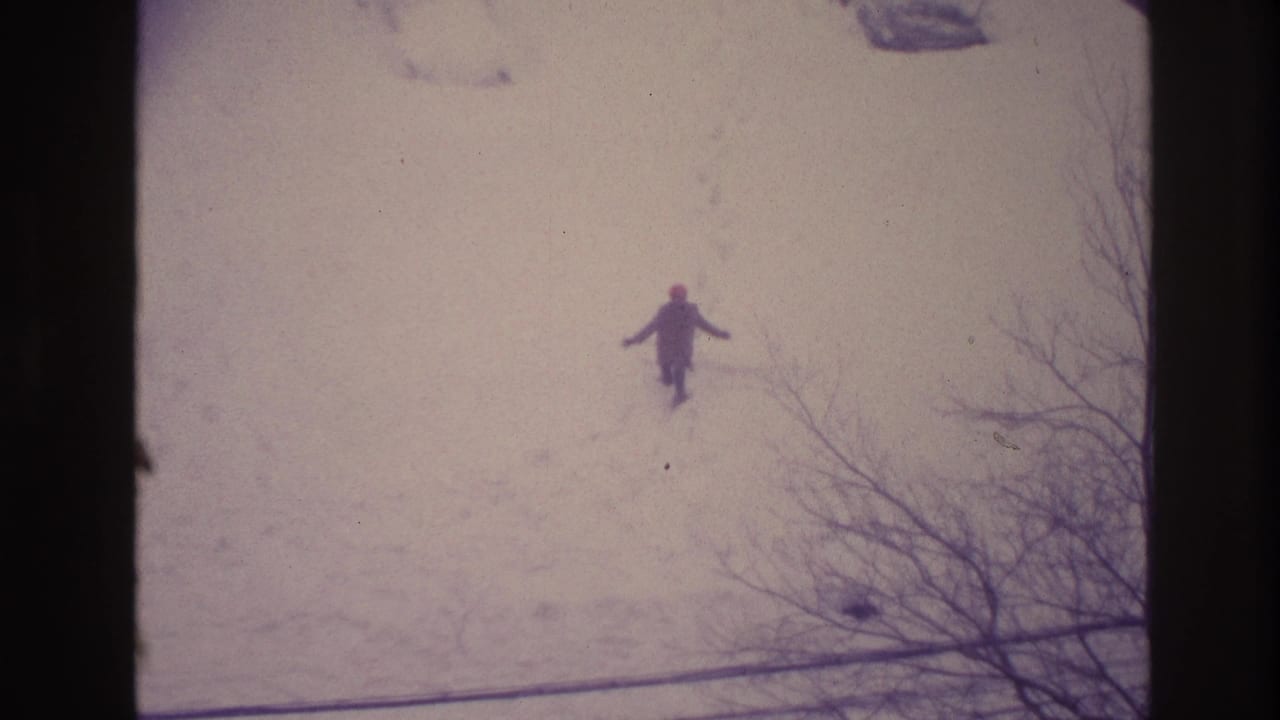 Distant figure stands or walks through snow with one arm raised, seen from above.
