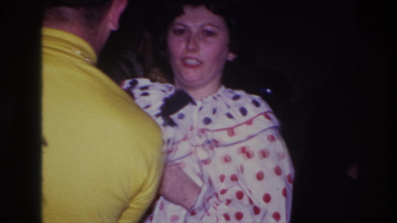 Ottawa 1967 frame of a woman in a white polka-dot clown costume speaking with a man in a bright yellow shirt in a dark room.