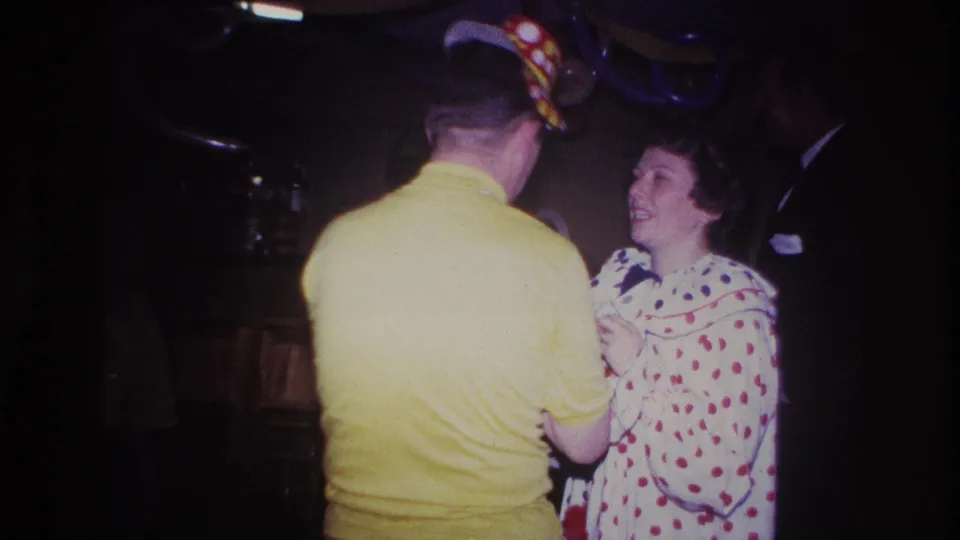 Ottawa 1967 frame with the clown-costumed woman smiling near the man in yellow, with dim furnishings and other guests barely visible.