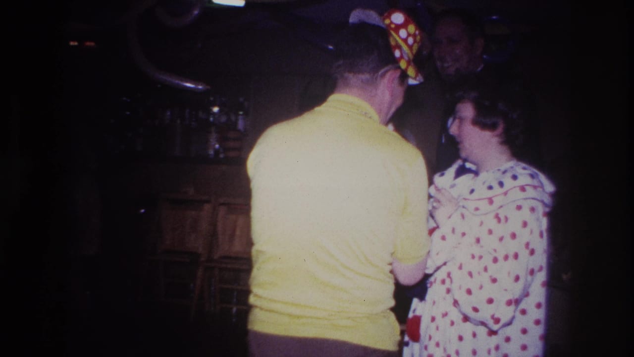 1967 Ottawa home-movie frame showing a man in a yellow shirt and party hat facing a woman in a white dress with red dots near a wood bar.