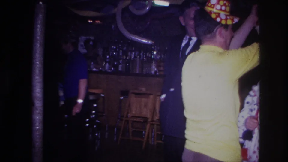 A 1967 Ottawa home-movie frame shows the man in yellow raising his arm near party guests, with bar stools and decorations in the background.