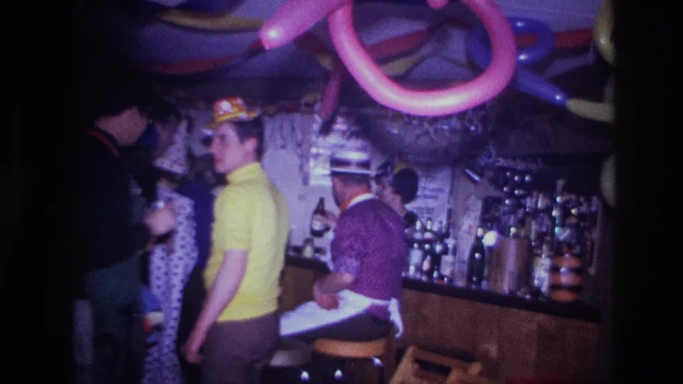 Ottawa 1967 home-movie frame showing a man in a yellow shirt facing the camera near adults, party hats, balloons, and a bar.