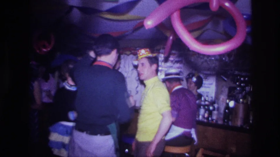 Ottawa 1967 frame showing party guests at a dim bar, including a man in a yellow shirt, paper hats, balloons, and bottles.