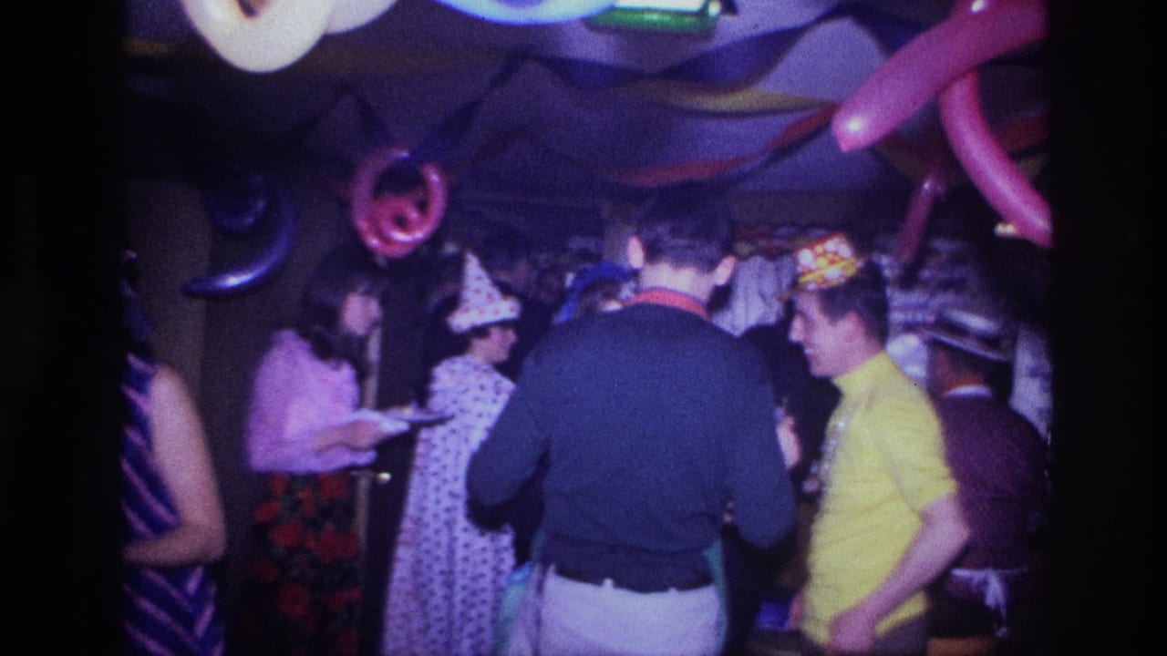 Party guests talk near balloons, streamers, and a table in a dim room.