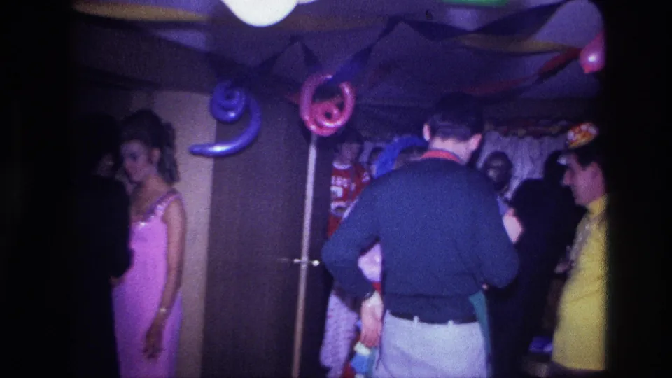 1967 Ottawa home-movie frame showing party guests near a doorway, including a woman in a pink dress and colorful hanging balloons.