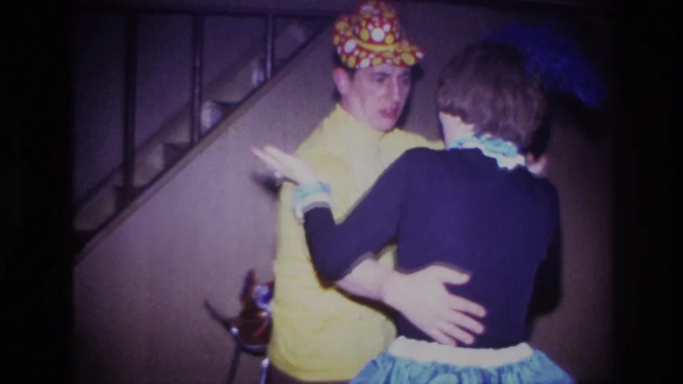 A woman in a blue ruffled skirt gestures while dancing with a man in a yellow costume near stairs in 1967 Ottawa footage.