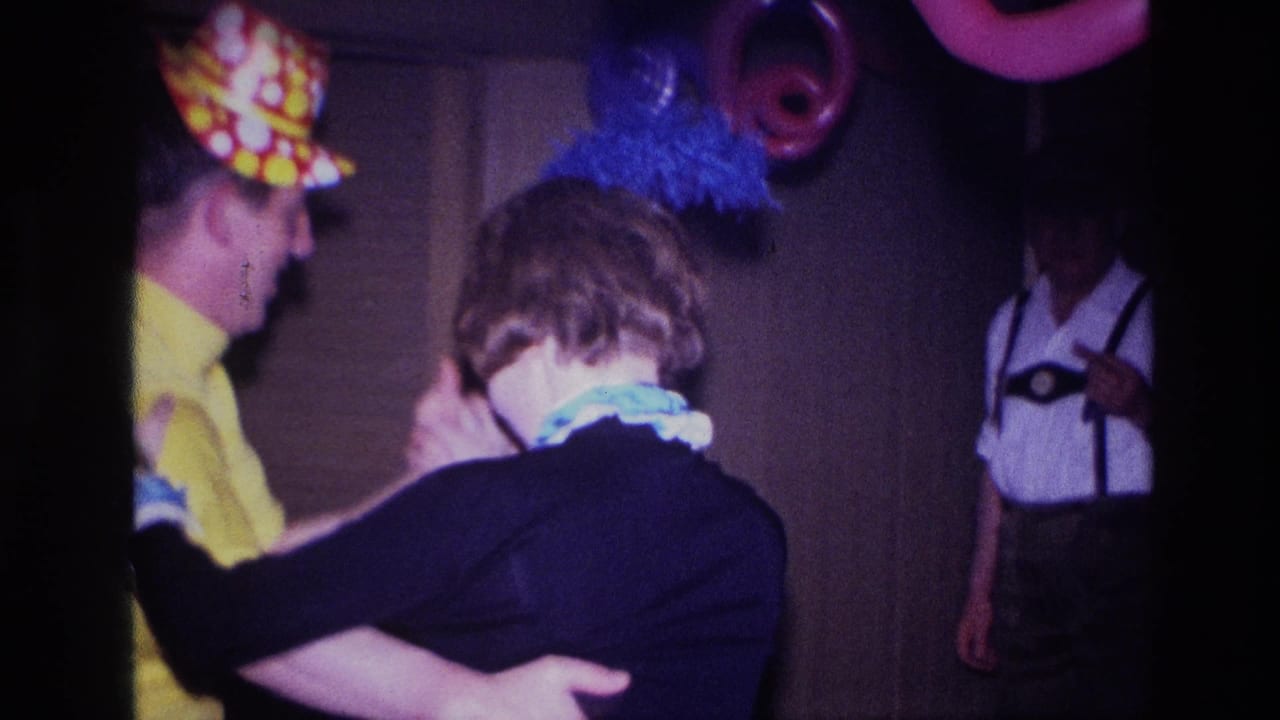 In a 1967 Ottawa home-movie frame, a couple dances in party hats as a guest in lederhosen-style dress watches from the dim room.