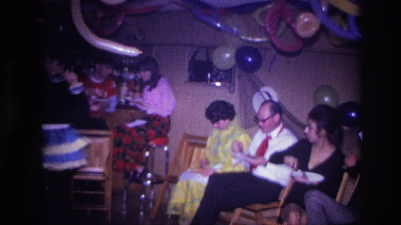 1967 Ottawa home-movie frame showing adults seated on wooden chairs at an indoor party with balloons, streamers, and paper plates.