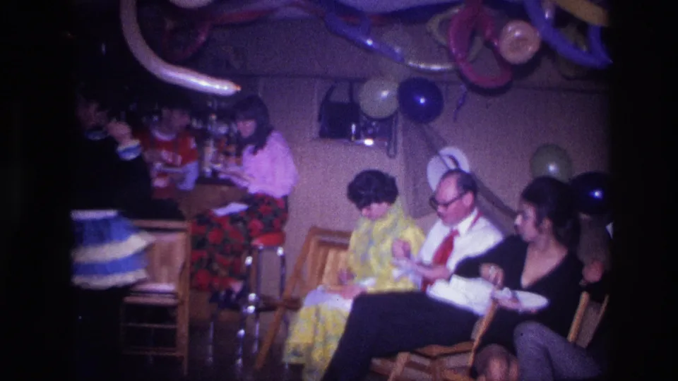 1967 Ottawa home-movie frame showing adults seated on wooden chairs at an indoor party with balloons, streamers, and paper plates.
