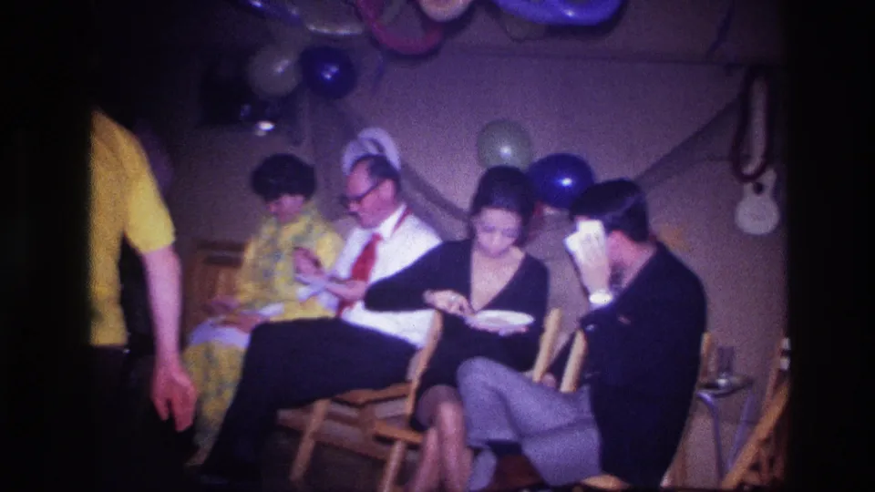 Home-movie frame from the Ottawa party with a guest passing near the camera as seated adults continue eating under balloons.