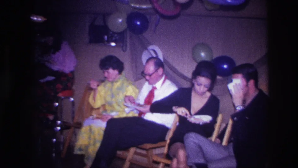 1967 Ottawa party frame showing adults seated closely together with plates, framed by balloons and streamers in a private interior.