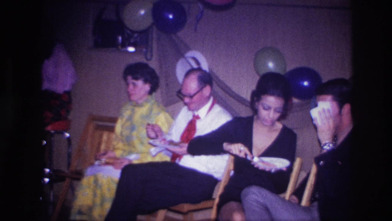 Adults seated in chairs eat from paper plates at a decorated indoor party.