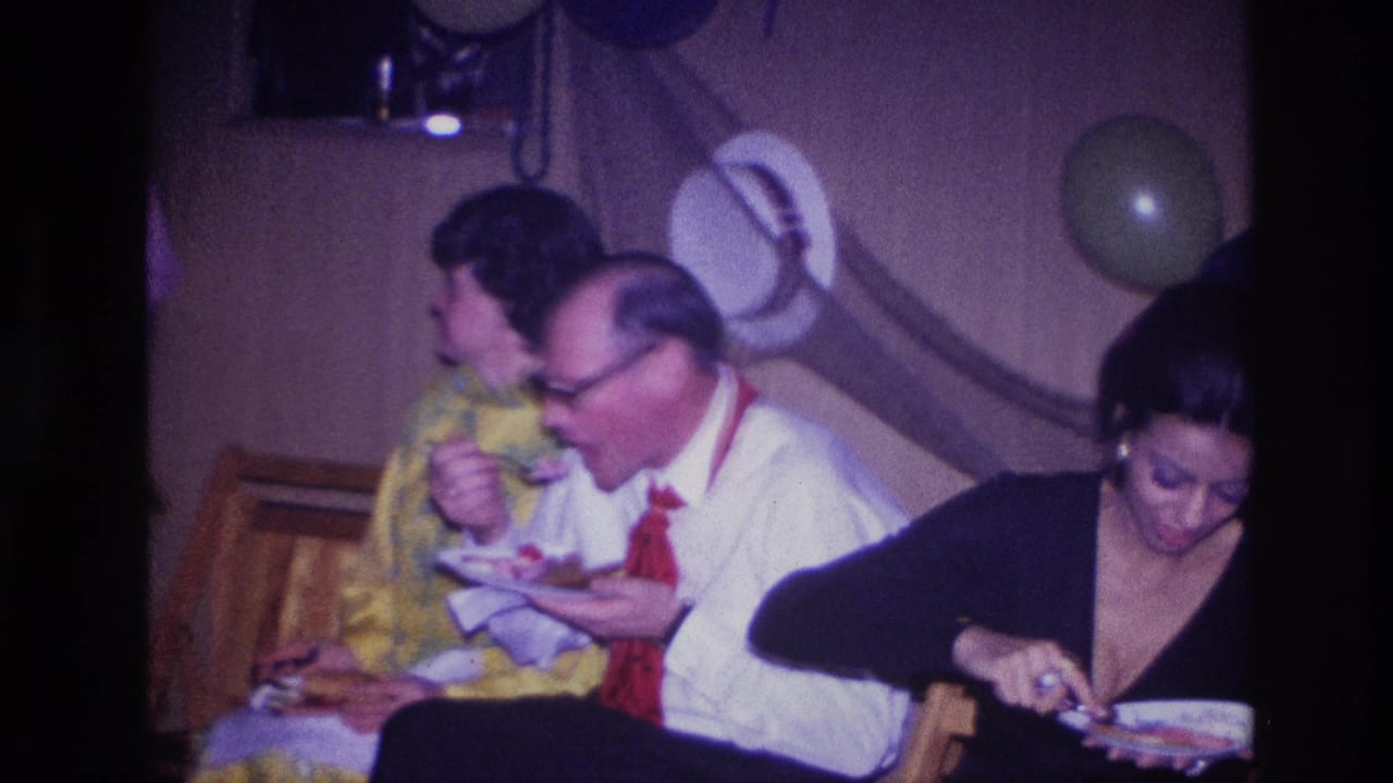 Adults eat from plates while balloons hang on the wall behind them.