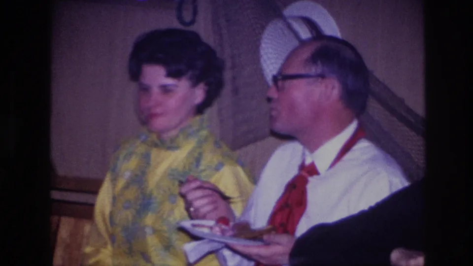 Close Ottawa 1967 home-movie frame of two adults at a party, with a woman in yellow and a man holding food on a plate.