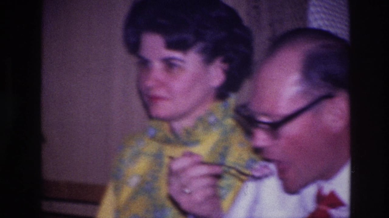 In a 1967 Ottawa home-movie frame, a woman in a yellow patterned dress sits beside a man with glasses eating from a plate.