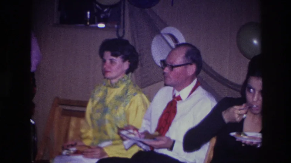 The frame widens to three adults seated indoors at the Ottawa gathering, including a woman eating from a plate near the camera.
