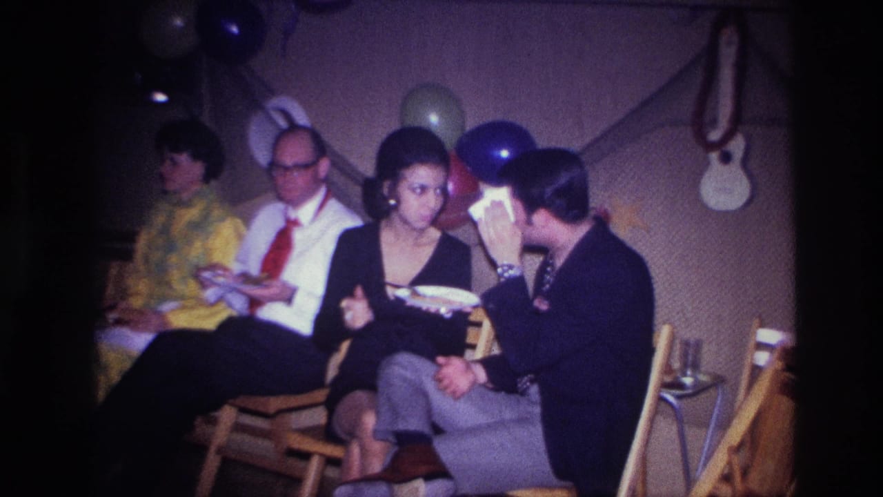 1967 Ottawa home-movie frame showing adults seated at a decorated indoor party, with guests eating and talking near balloons and streamers.